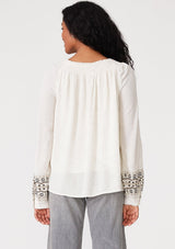 [Color: Natural] A back facing image of a brunette model wearing a bohemian blouse with embroidered details. With voluminous long sleeves, a split v neckline with tassel ties, a smocked neckline, and a relaxed fit. 