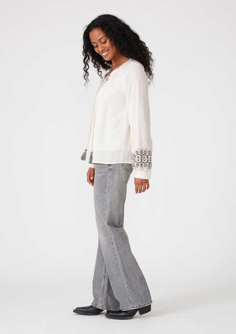 [Color: Natural] A side facing image of a brunette model wearing a bohemian blouse with embroidered details. With voluminous long sleeves, a split v neckline with tassel ties, a smocked neckline, and a relaxed fit. 