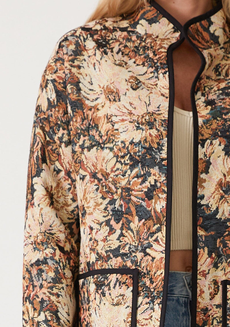 [Color: Black/Natural] A close up front facing image of a blonde model wearing a bohemian jacket in a black and natural floral tapestry. With long sleeves, a dropped shoulder, an open front, and side patch pockets.