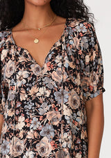 [Color: Black/Dusty Blue] A close up front facing image of a brunette model wearing a short puff sleeve blouse in a black and dusty blue floral print. With a split v neckline with tassel ties and a self covered button front. 