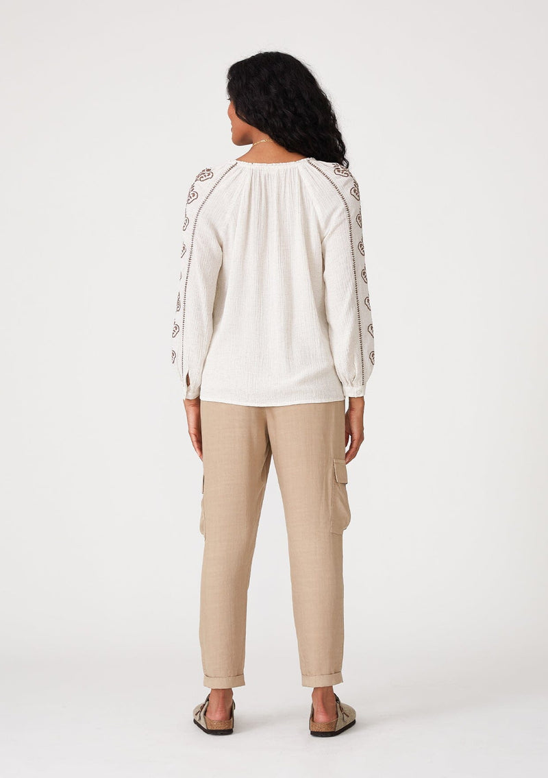 [Color: Natural/Taupe] A back facing image of a brunette model wearing a bohemian off white blouse with embroidered detail. With voluminous long sleeves, a v neckline, and a relaxed fit. 
