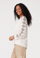 [Color: Natural/Taupe] A side facing image of a brunette model wearing a bohemian off white blouse with embroidered detail. With voluminous long sleeves, a v neckline, and a relaxed fit. 