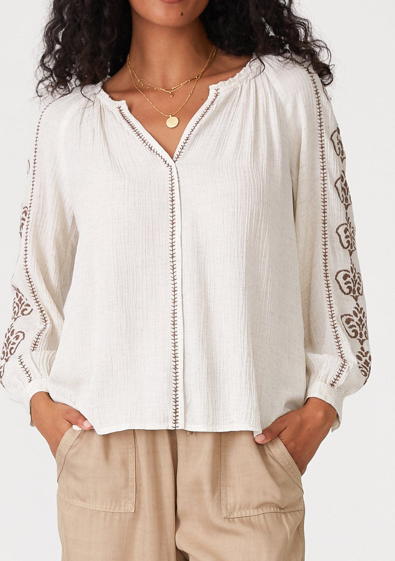 [Color: Natural/Taupe] A close up front facing image of a brunette model wearing a bohemian off white blouse with embroidered detail. With voluminous long sleeves, a v neckline, and a relaxed fit. 