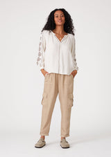 [Color: Natural/Taupe] A full body front facing image of a brunette model wearing a bohemian off white blouse with embroidered detail. With voluminous long sleeves, a v neckline, and a relaxed fit. 