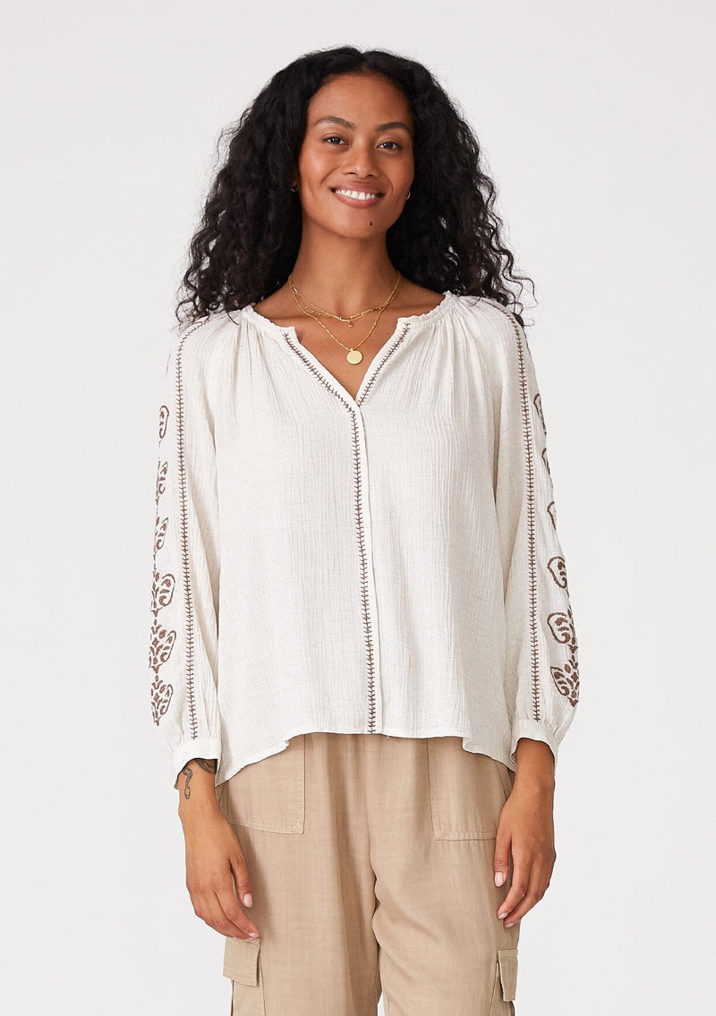 [Color: Natural/Taupe] A front facing image of a brunette model wearing a bohemian off white blouse with embroidered detail. With voluminous long sleeves, a v neckline, and a relaxed fit. 