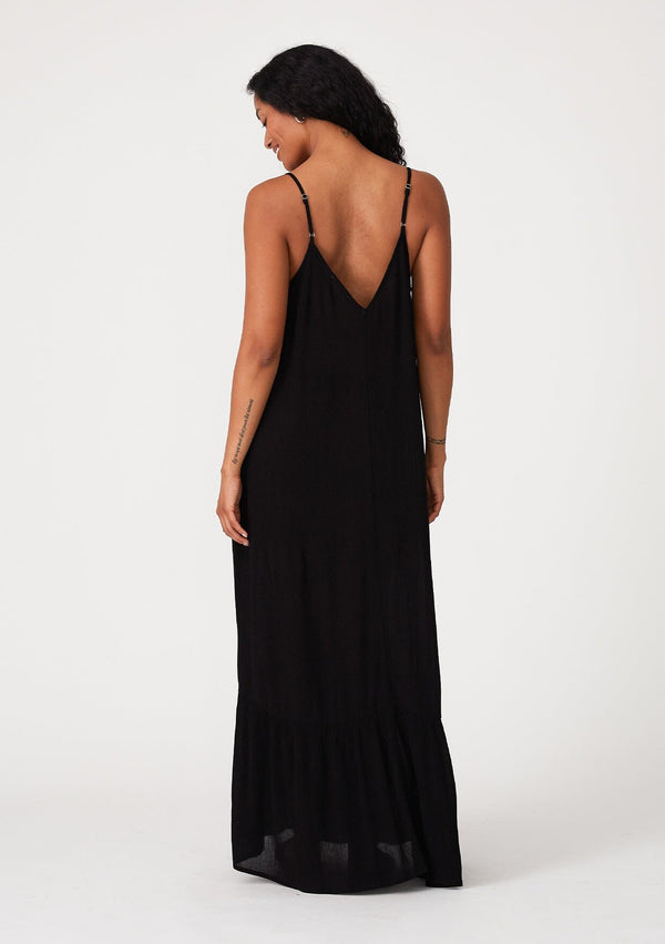 [Color: Black] A back facing image of a brunette model wearing a simple flowy sleeveless maxi tank dress in a black crinkle rayon. With a v neckline in front and back, adjustable spaghetti straps, and a tiered skirt.