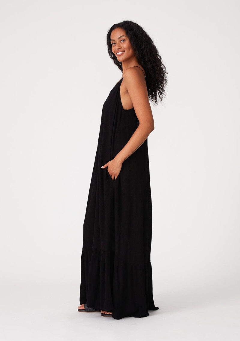 [Color: Black] A side facing image of a brunette model wearing a simple flowy sleeveless maxi tank dress in a black crinkle rayon. With a v neckline in front and back, adjustable spaghetti straps, and a tiered skirt.