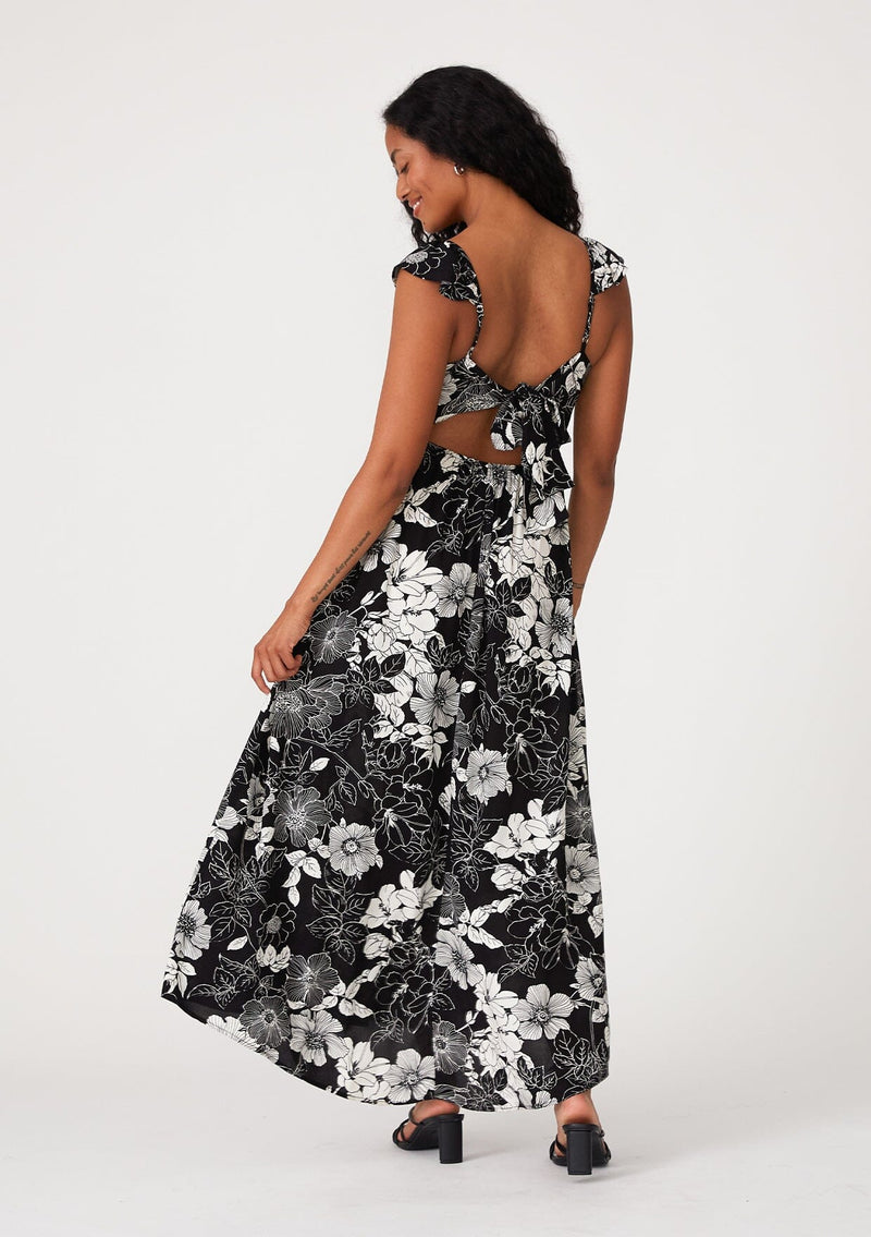 [Color: Black/White] A back facing image of a brunette model wearing a pretty bohemian maxi dress in a black and white floral print. With short flutter cap sleeves, a scooped neckline, side pockets, adjustable spaghetti straps, and an adjustable tie back detail. 