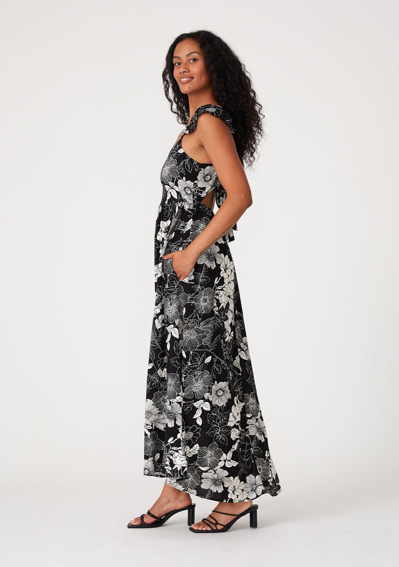 [Color: Black/White] A side facing image of a brunette model wearing a pretty bohemian maxi dress in a black and white floral print. With short flutter cap sleeves, a scooped neckline, side pockets, adjustable spaghetti straps, and an adjustable tie back detail. 