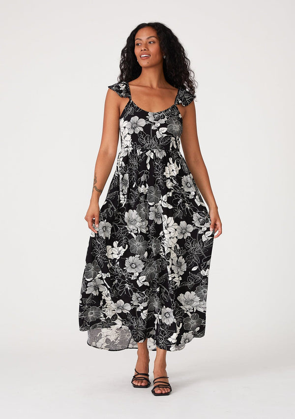 [Color: Black/White] A front facing image of a brunette model wearing a pretty bohemian maxi dress in a black and white floral print. With short flutter cap sleeves, a scooped neckline, side pockets, adjustable spaghetti straps, and an adjustable tie back detail. 