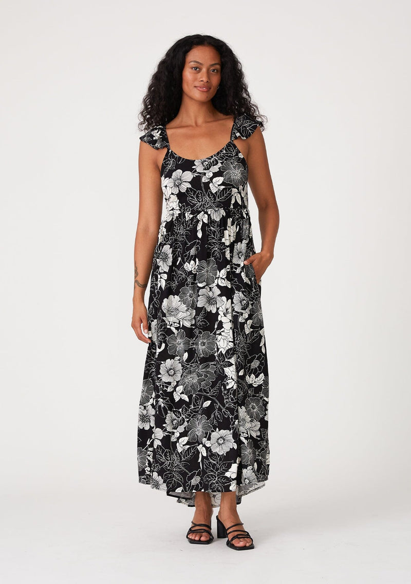 [Color: Black/White] A full body front facing image of a brunette model wearing a pretty bohemian maxi dress in a black and white floral print. With short flutter cap sleeves, a scooped neckline, side pockets, adjustable spaghetti straps, and an adjustable tie back detail. 