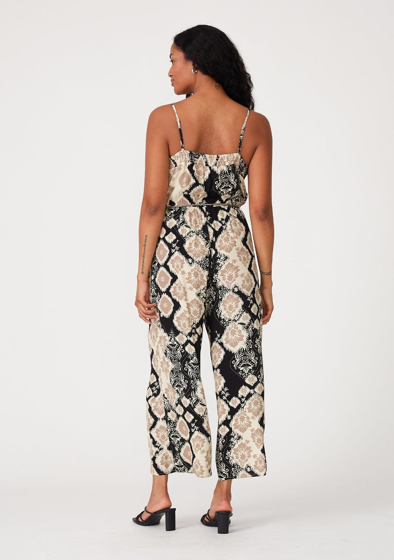 [Color: Black/Khaki] A back facing image of a brunette model wearing a sleeveless jumpsuit in a black and beige bohemian print. With adjustable spaghetti straps, a scoop neckline, side pockets, a long wide leg, and a beaded braided belt. 
