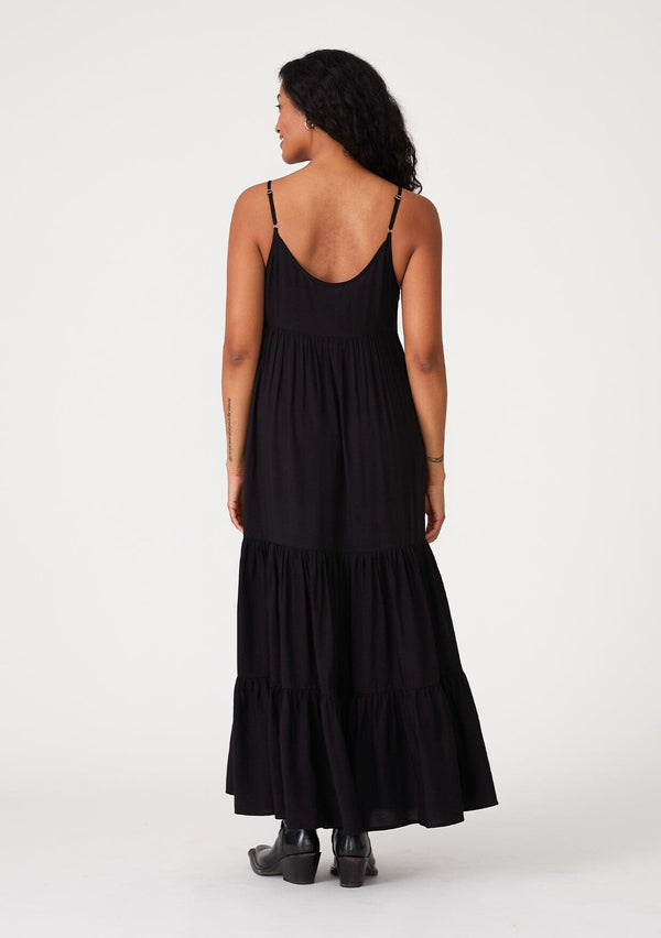 [Color: Black] A back facing image of a brunette model wearing a black sleeveless bohemian maxi dress. With a contrast embroidered scallop trim, adjustable spaghetti straps, a tiered long skirt, side pockets, and a self covered button front. 