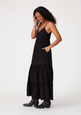 [Color: Black] A side facing image of a brunette model wearing a black sleeveless bohemian maxi dress. With a contrast embroidered scallop trim, adjustable spaghetti straps, a tiered long skirt, side pockets, and a self covered button front. 