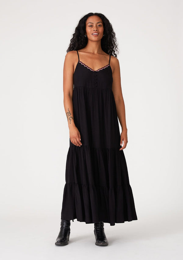 [Color: Black] A front facing image of a brunette model wearing a black sleeveless bohemian maxi dress. With a contrast embroidered scallop trim, adjustable spaghetti straps, a tiered long skirt, side pockets, and a self covered button front. 