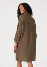 [Color: Military] A back facing image of a brunette model wearing an olive green relaxed fit shirt dress. With long sleeves, a button front, a high low hemline, a collared neckline, and front flap pockets. 