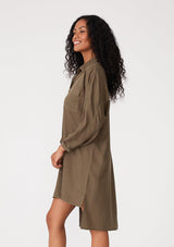[Color: Military] A side facing image of a brunette model wearing an olive green relaxed fit shirt dress. With long sleeves, a button front, a high low hemline, a collared neckline, and front flap pockets. 
