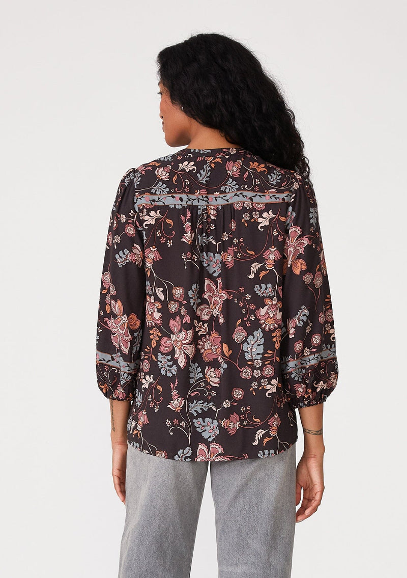 [Color: Brown/Dusty Lilac] A back facing image of a brunette model wearing a bohemian fall blouse in a brown and dusty purple floral print. With three quarter length sleeves, a self covered button front, a split v neckline with tassel ties, and a relaxed fit. 