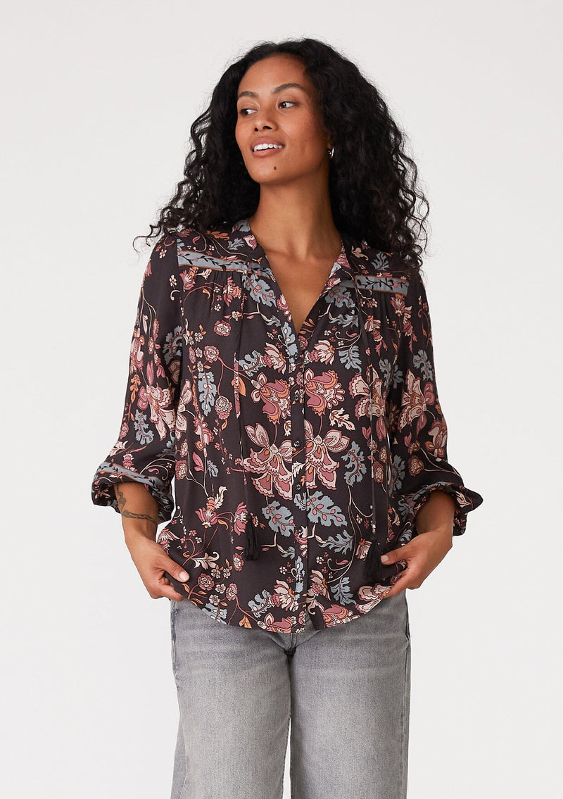 [Color: Brown/Dusty Lilac] A front facing image of a brunette model wearing a bohemian fall blouse in a brown and dusty purple floral print. With three quarter length sleeves, a self covered button front, a split v neckline with tassel ties, and a relaxed fit. 