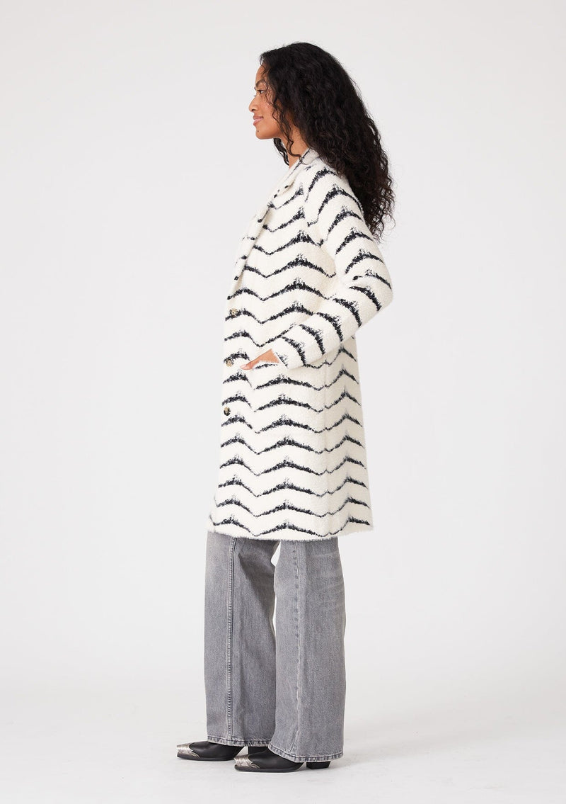 [Color: Cream/Black] A side facing image of a brunette model wearing a soft and fuzzy sweater coat in a white and black chevron design. With a snap button front, side pockets, and a classic notched lapel.