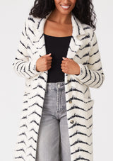 [Color: Cream/Black] A close up front facing image of a brunette model wearing a soft and fuzzy sweater coat in a white and black chevron design. With a snap button front, side pockets, and a classic notched lapel.