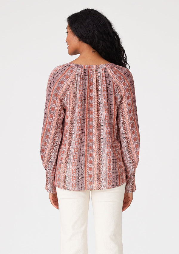[Color: Cinnamon/Coral] A back facing image of a brunette model wearing a bohemian fall blouse in a pink print. With long sleeves, a loop button front, a v neckline, and tapered sleeves. 