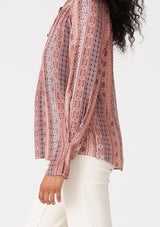 [Color: Cinnamon/Coral] A close up side facing image of a brunette model wearing a bohemian fall blouse in a pink print. With long sleeves, a loop button front, a v neckline, and tapered sleeves. 