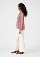 [Color: Cinnamon/Coral] A side facing image of a brunette model wearing a bohemian fall blouse in a pink print. With long sleeves, a loop button front, a v neckline, and tapered sleeves. 