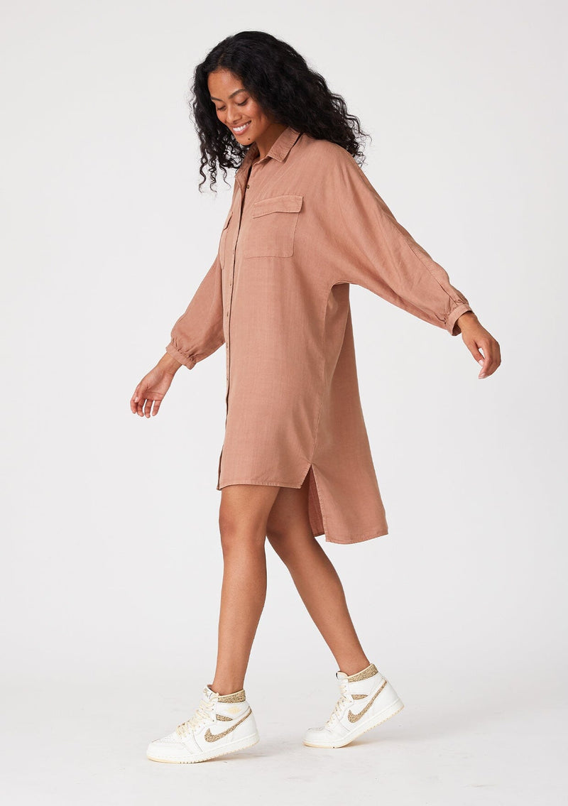 [Color: Clay] A side facing image of a brunette model wearing a pink relaxed fit shirt dress. With long sleeves, a button front, a high low hemline, a collared neckline, and front flap pockets. 