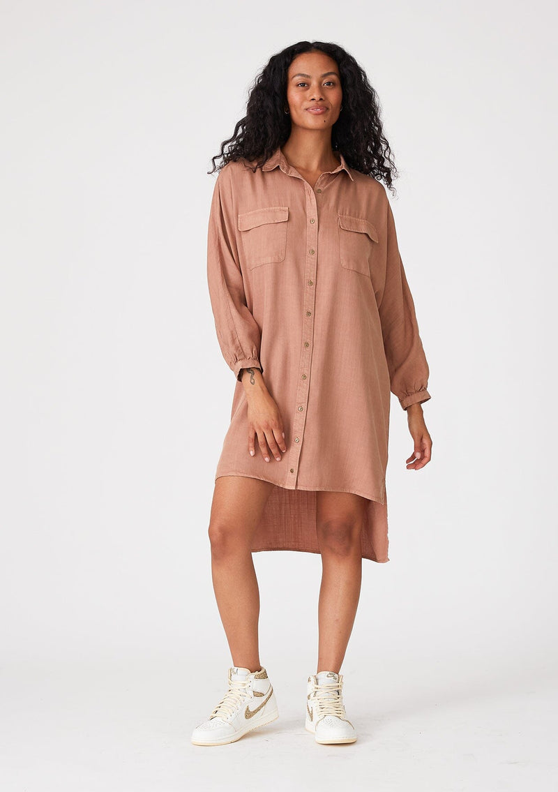 [Color: Clay] A front facing image of a brunette model wearing a pink relaxed fit shirt dress. With long sleeves, a button front, a high low hemline, a collared neckline, and front flap pockets. 