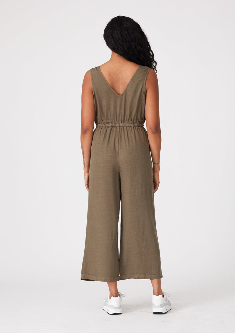 [Color: Military] A back facing image of a brunette model wearing an olive green sleeveless jumpsuit with a cropped wide leg, a surplice v neckline, side pockets, and a drawstring tie waist. 