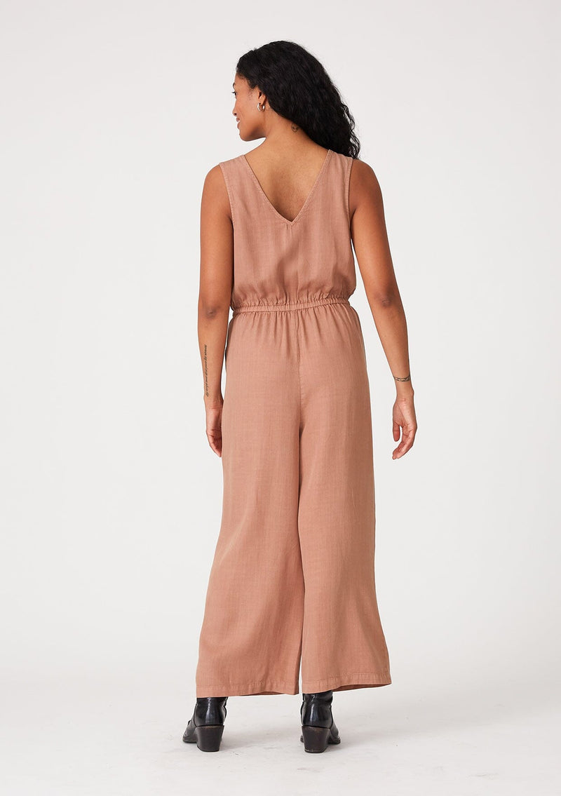 [Color: Clay] A back facing image of a brunette model wearing a pink sleeveless jumpsuit with a cropped wide leg, a surplice v neckline, side pockets, and a drawstring tie waist. 