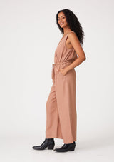 [Color: Clay] A side facing image of a brunette model wearing a pink sleeveless jumpsuit with a cropped wide leg, a surplice v neckline, side pockets, and a drawstring tie waist. 