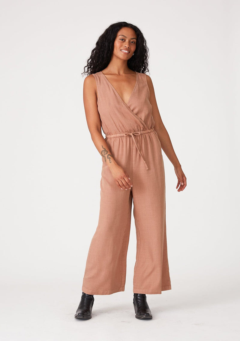 [Color: Clay] A front facing image of a brunette model wearing a pink sleeveless jumpsuit with a cropped wide leg, a surplice v neckline, side pockets, and a drawstring tie waist. 