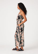 [Color: Black/Dusty Rose] A side facing image of a brunette model wearing a sleeveless jumpsuit in a black and pink bohemian print. With adjustable spaghetti straps, a scoop neckline, side pockets, a long wide leg, and a beaded braided belt. 