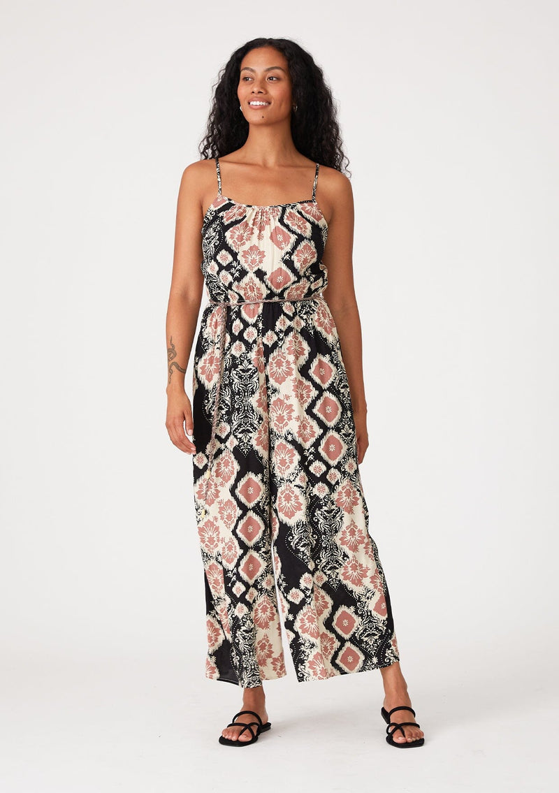[Color: Black/Dusty Rose] A front facing image of a brunette model wearing a sleeveless jumpsuit in a black and pink bohemian print. With adjustable spaghetti straps, a scoop neckline, side pockets, a long wide leg, and a beaded braided belt. 