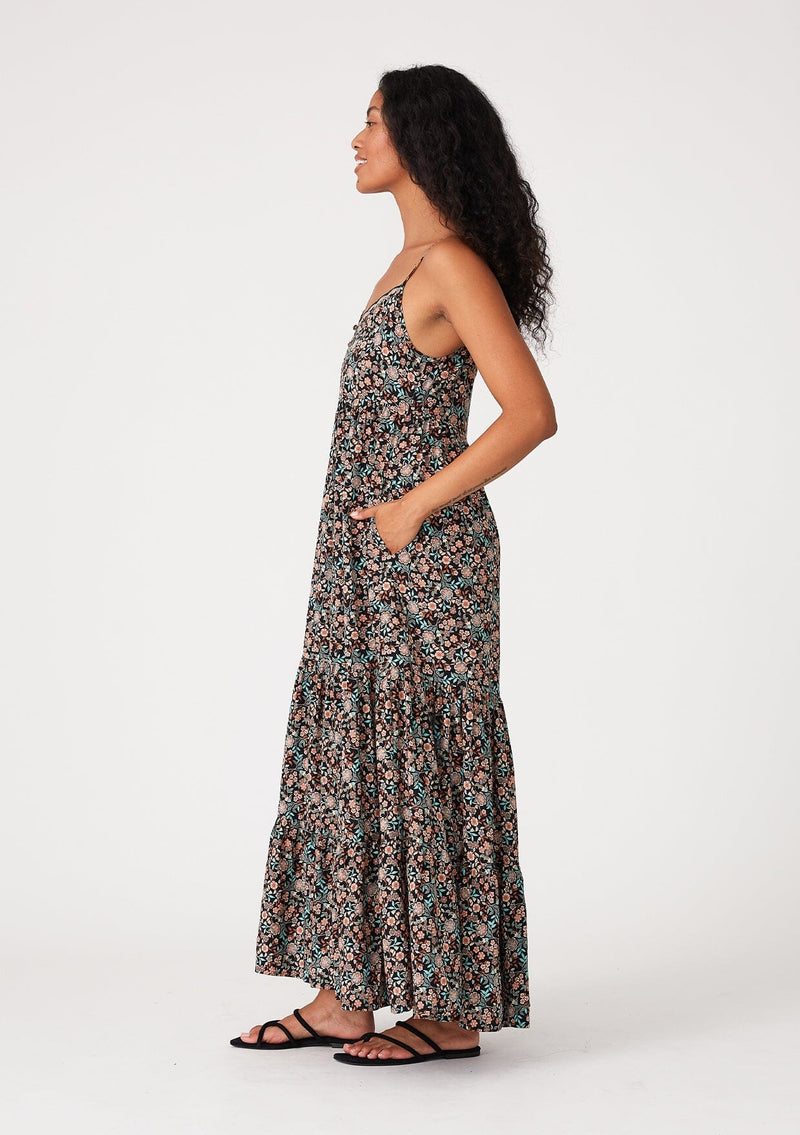 [Color: Black/Teal] A side facing image of a brunette model wearing a bohemian maxi dress in a black and teal floral print. With adjustable spaghetti straps, a scalloped trim v neckline with contrast thread details, a tiered flowy silhouette, side pockets, and an empire waist.