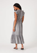 [Color: Grey] A back facing image of a brunette model wearing a classic bohemian maxi dress in soft grey. With short flutter cap sleeves, a v neckline, a self covered button front a tiered skirt, a half smocked elastic waist at the back, and eyelet lace details throughout. 