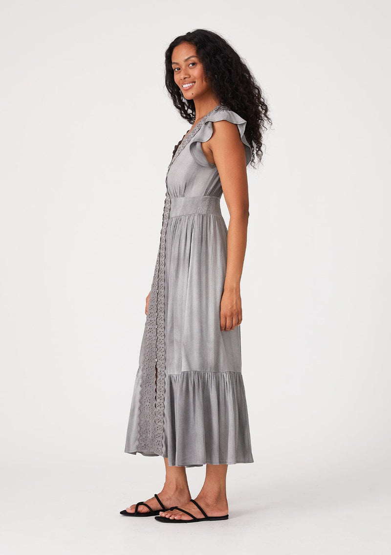 [Color: Grey] A side facing image of a brunette model wearing a classic bohemian maxi dress in soft grey. With short flutter cap sleeves, a v neckline, a self covered button front a tiered skirt, a half smocked elastic waist at the back, and eyelet lace details throughout. 