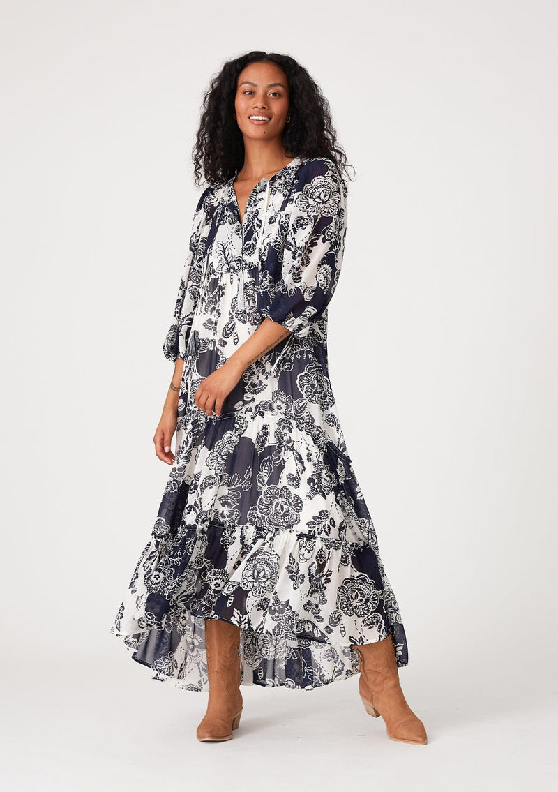 [Color: Natural/Navy] A front facing image of a brunette model wearing a flowy bohemian maxi dress in a blue and white floral print. With three quarter length puff sleeves, tie cuff details, a tiered high low skirt, a button front, ruffle details, and tassel neck ties. 