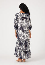 [Color: Natural/Navy] A back facing image of a brunette model wearing a flowy bohemian maxi dress in a blue and white floral print. With three quarter length puff sleeves, tie cuff details, a tiered high low skirt, a button front, ruffle details, and tassel neck ties. 
