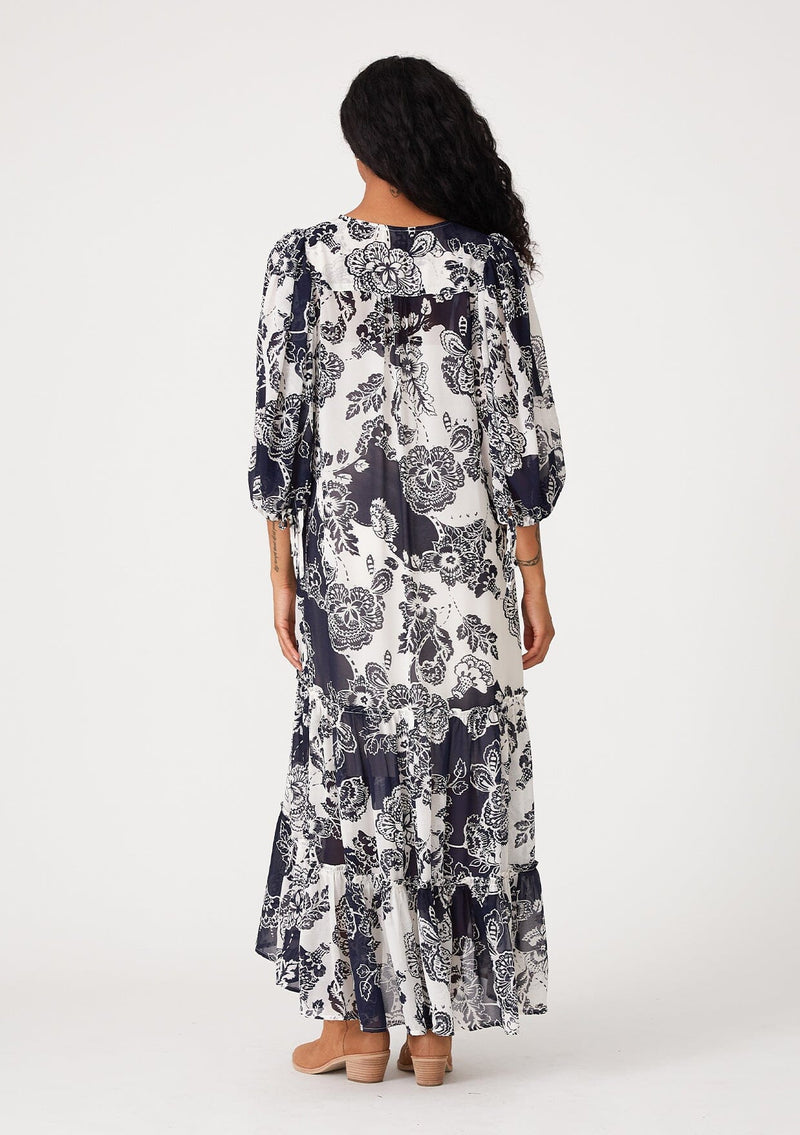 [Color: Natural/Navy] A back facing image of a brunette model wearing a flowy bohemian maxi dress in a blue and white floral print. With three quarter length puff sleeves, tie cuff details, a tiered high low skirt, a button front, ruffle details, and tassel neck ties. 