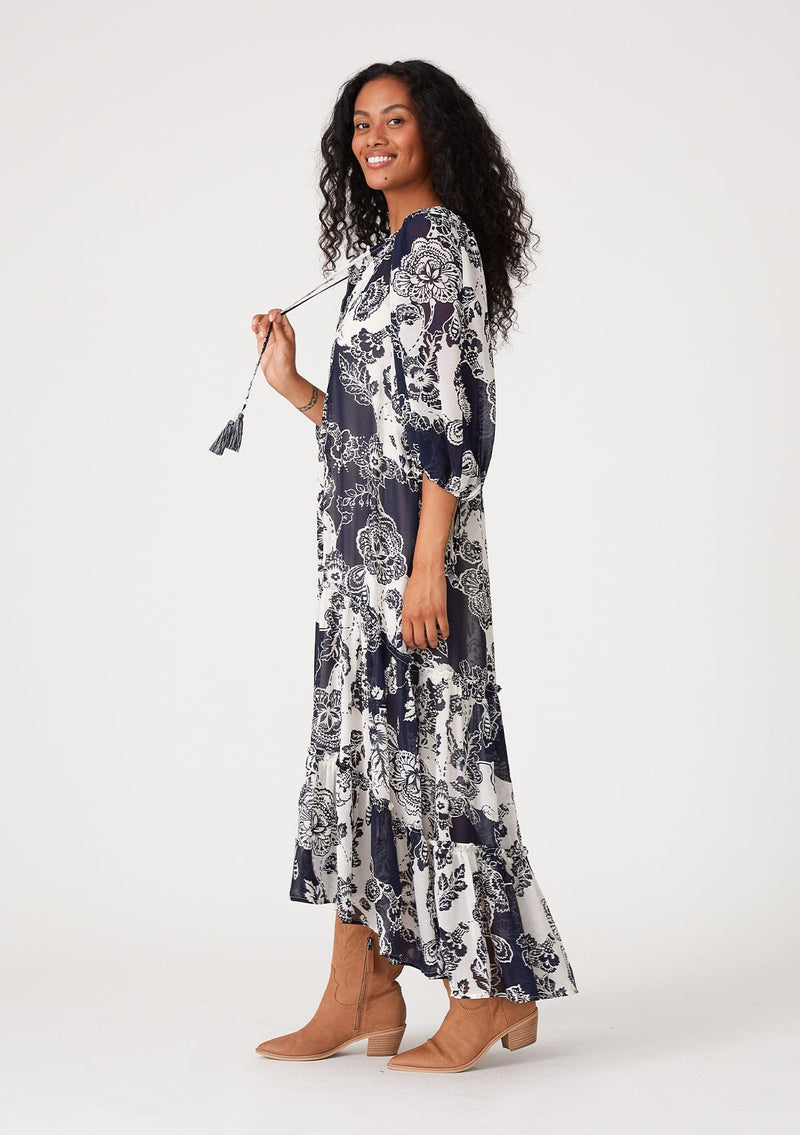 [Color: Natural/Navy] A side facing image of a brunette model wearing a flowy bohemian maxi dress in a blue and white floral print. With three quarter length puff sleeves, tie cuff details, a tiered high low skirt, a button front, ruffle details, and tassel neck ties. 