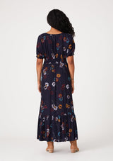 [Color: Navy/Mustard] A back facing image of a brunette model wearing a bohemian fall maxi dress in a navy blue and mustard yellow floral print. With short puff sleeves, a v neckline, a long flowy tiered skirt, and a side slit.