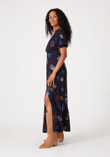 [Color: Navy/Mustard] A side facing image of a brunette model wearing a bohemian fall maxi dress in a navy blue and mustard yellow floral print. With short puff sleeves, a v neckline, a long flowy tiered skirt, and a side slit.