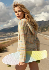[Color: Sherbert] A full body side facing image of a blonde model at the beach wearing a multi color space dye knit sweater. A bohemian knit hoodie with long sleeves, a kangaroo front pocket, dropped shoulders, and a drawstring hood.