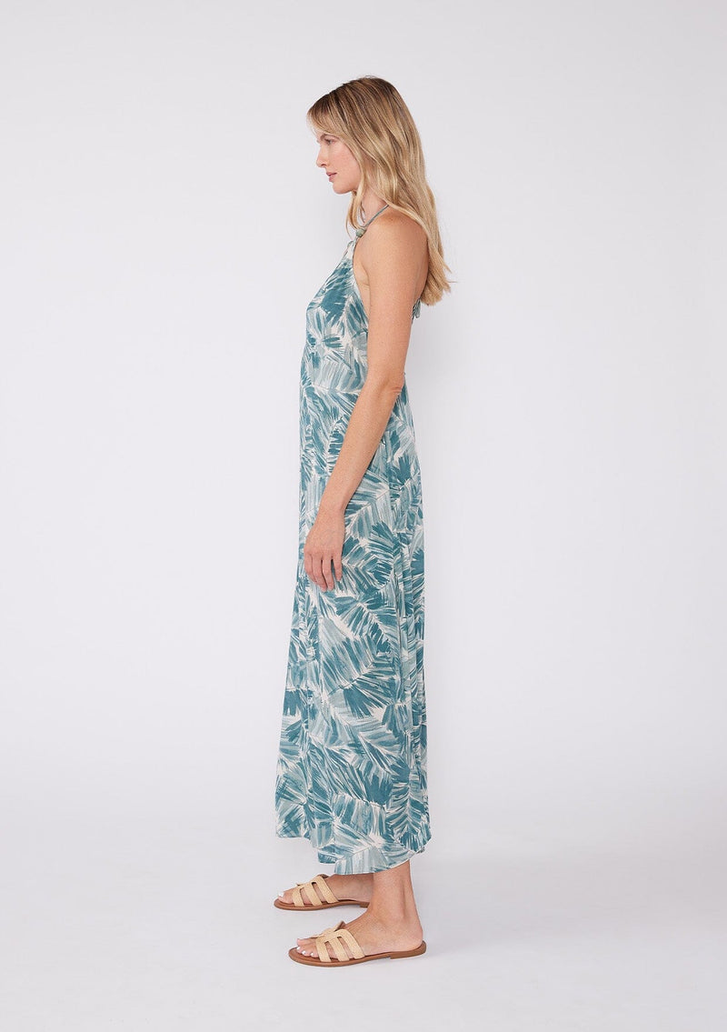 [Color: Natural/Green] A side facing image of a blonde model wearing a sleeveless summer maxi dress in a tropical green palm leaf print. With a v neckline, spaghetti straps with bead accents, a strappy back with adjustable tie, and a flowy silhouette. 