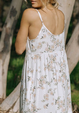 [Color: Ivory/Dusty Coral] A front facing image of a brunette model wearing a pretty bohemian sleeveless maxi dress in a pink and white floral print. With adjustable spaghetti straps, a scalloped trimmed v neckline, a decorative self covered button front top, a tiered flowy skirt, and an empire waist.