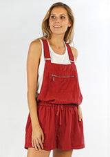 [Color: Brick] A front facing image of a blonde model wearing a red overall romper over a tank top with open sides, a front zipper details, drawstring waist, and side pockets. 