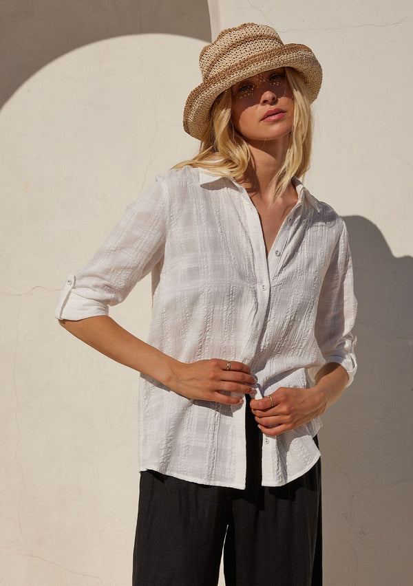 [Color: White] A front facing image of a blonde model standing outside wearing a classic bohemian white cotton relaxed shirt in a textured plaid seersucker fabric. With long sleeves, a button tab closure at the sleeve, a collared neckline, and a self covered button front. 